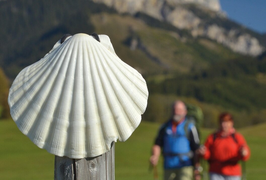 Jakobsmuschel auf Holzpfosten, Wanderer im Hintergrund, Berglandschaft
