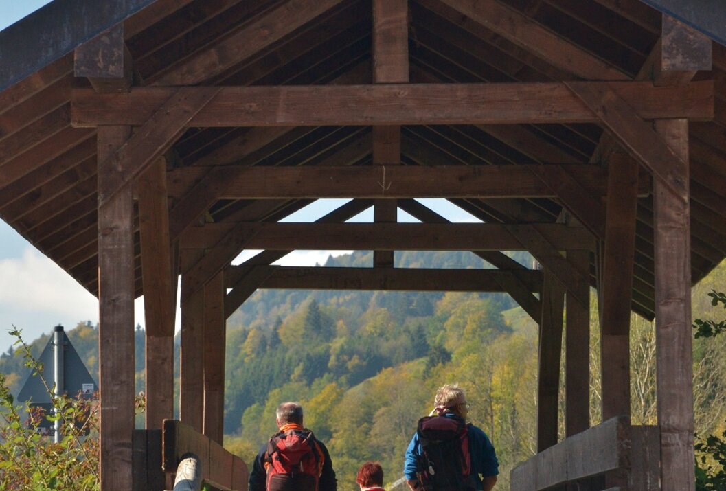 Wandernde Familie auf Holzbrücke mit Dach, im Hintergrund bewaldete Hügel