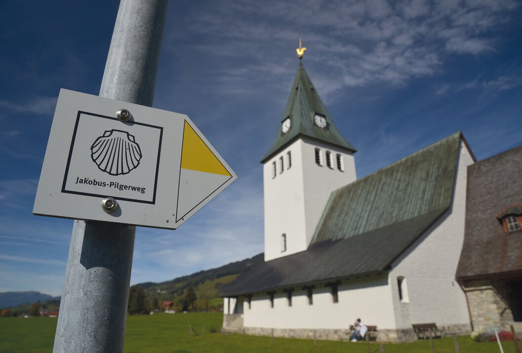 Jakobus-Pilgerweg Schild vor Kirche mit grünem Kirchturm und gelbem Wetterhahn