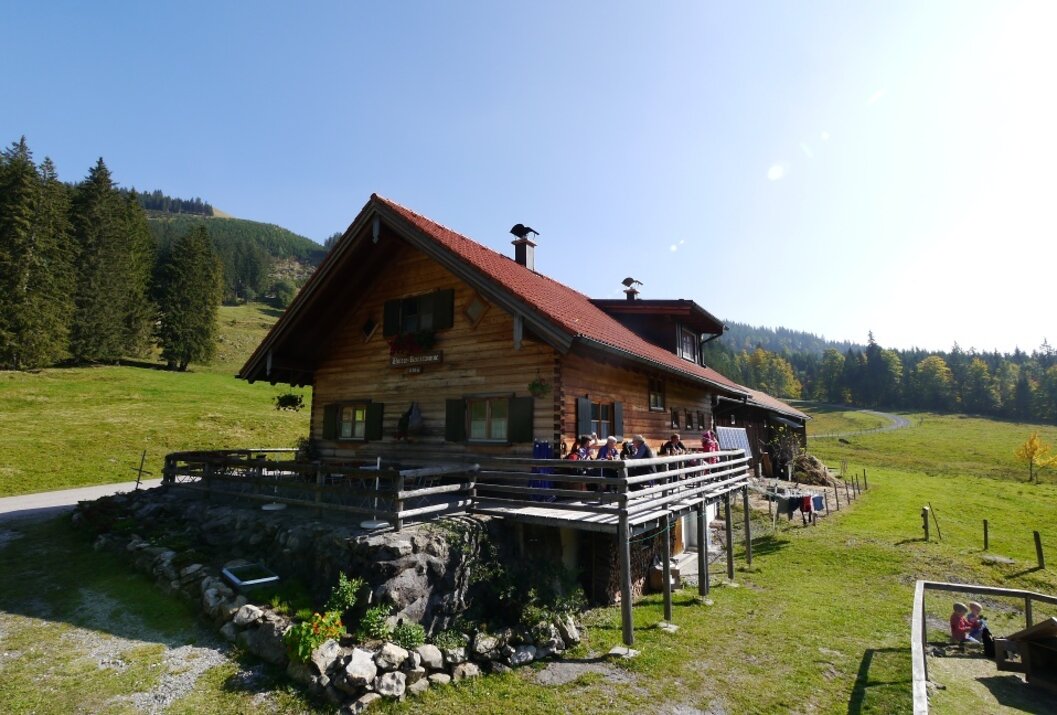 Holzhütte mit rotem Dach auf einer Wiese vor bewaldeten Hügeln