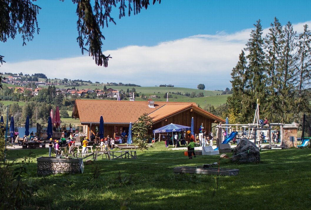 Hütte mit Spielplatz auf grüner Wiese vor Hügellandschaft unter blauem Himmel