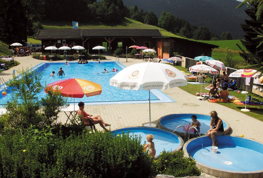 Freibad mit Schwimmbecken, Kinderbecken, Liegewiese, Sonnenschirmen und bewaldeten Bergen im Hintergrund