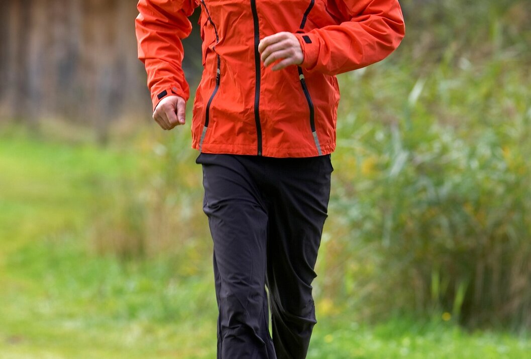 Mann in orangefarbener Jacke und dunkler Hose joggt im Grünen