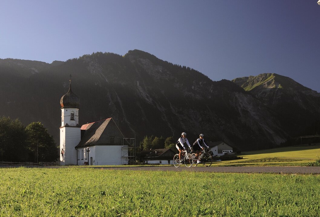 Zwei Radfahrer vor Kirche und Berglandschaft