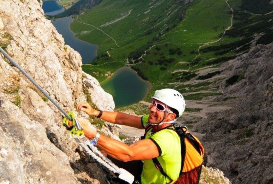 Bergsteiger am Felsen mit Seil, im Hintergrund Berglandschaft mit See