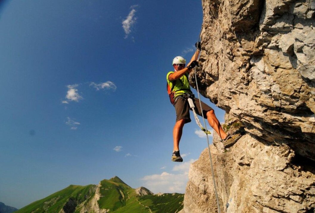 Kletterer am Felsen mit Seil und Helm vor blauem Himmel und Berglandschaft
