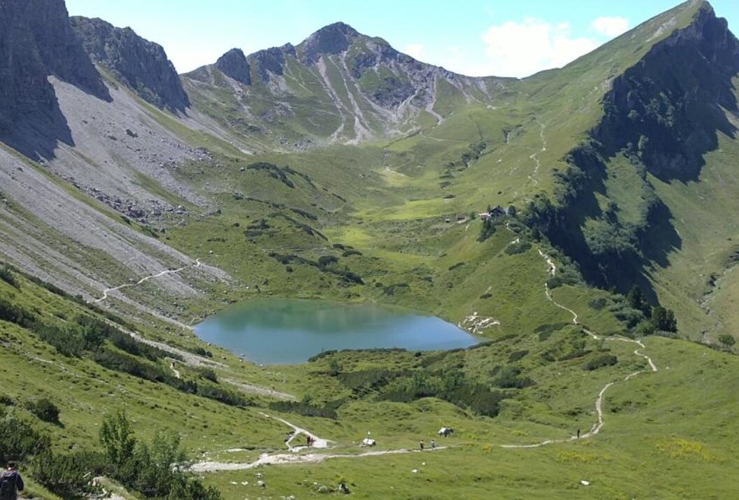 Bergsee eingebettet in grüne Hügellandschaft mit Wanderwegen und Berggipfel im Hintergrund
