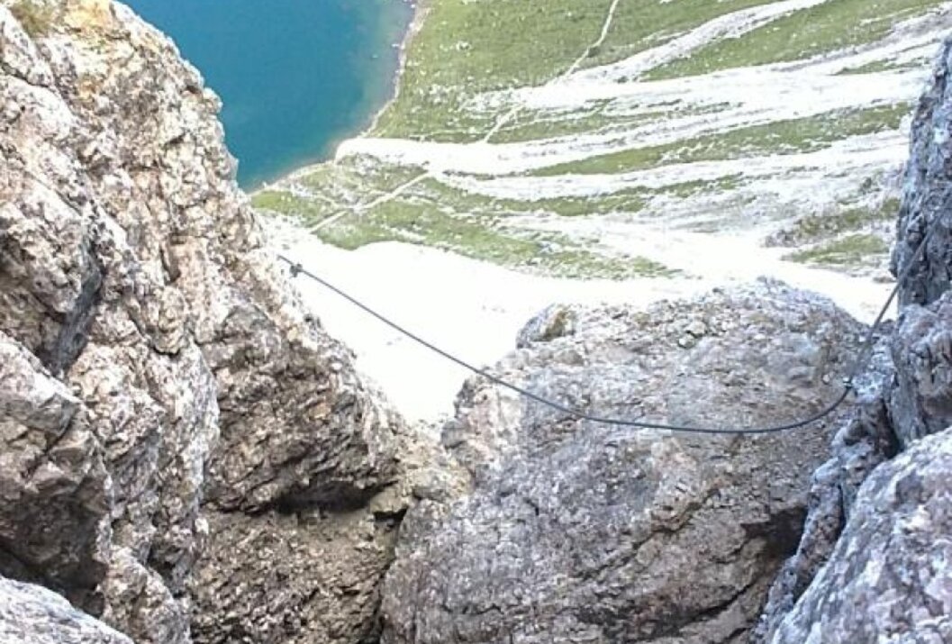 Felsiger Bergpfad mit Drahtseil, im Hintergrund ein blauer Bergsee und grüne Wiesen
