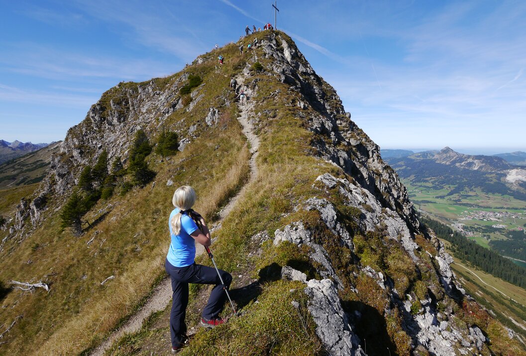 Frau blickt auf Bergpfad mit Gipfelkreuz und Wanderern