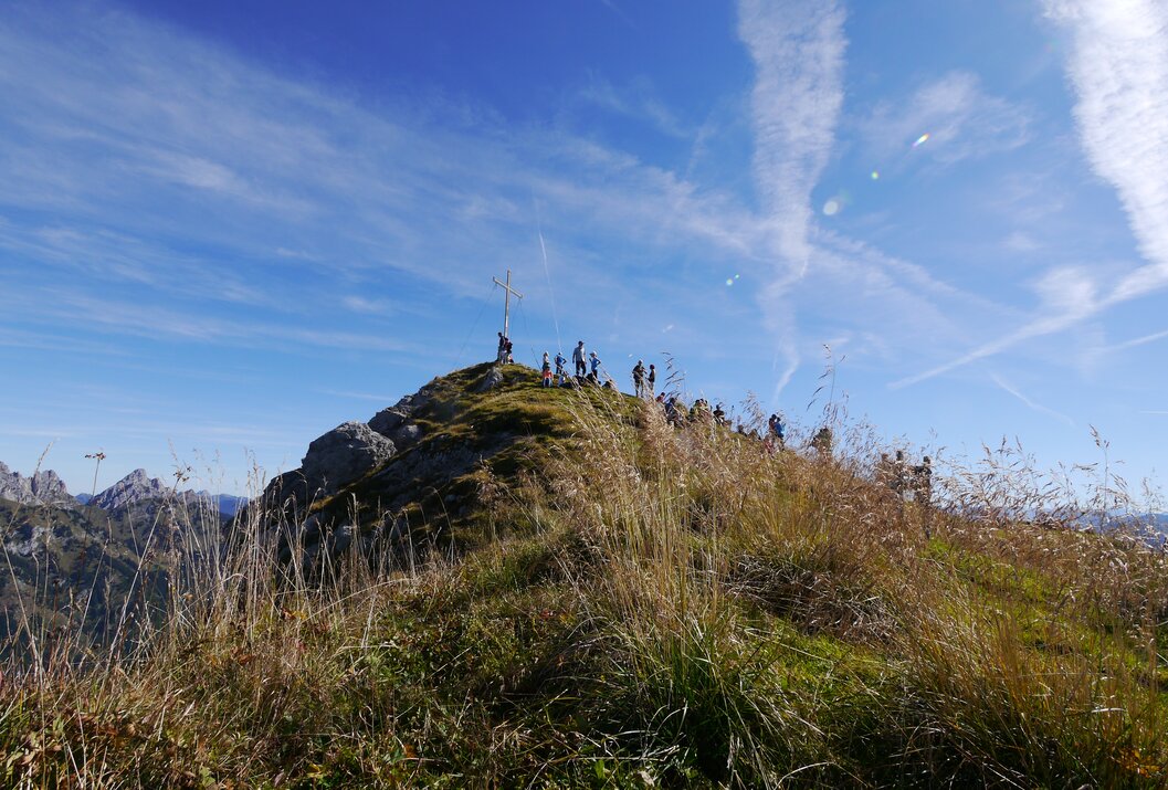 Bergspitze mit Gipfelkreuz, Menschengruppe und Gras im Vordergrund unter blauem Himmel