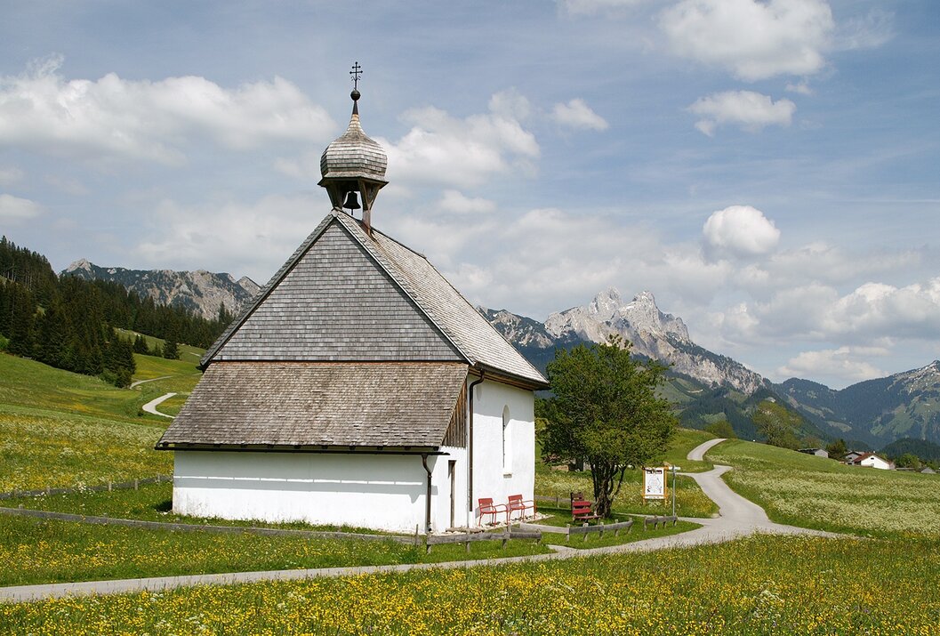 Kleine weiße Kapelle auf grüner Wiese, im Hintergrund Berglandschaft unter blauem Himmel
