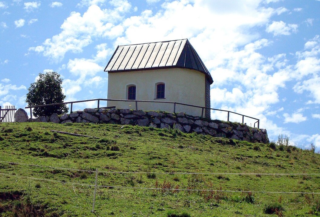 Kleine Kapelle auf einem Hügel mit Steinmauer und Holzzaun vor blauem Himmel mit Wolken
