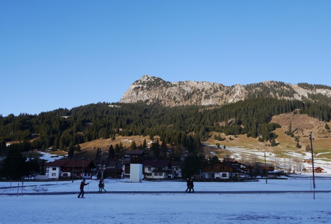 Winterlandschaft mit schneebedecktem Feld, Häusern und bewaldetem Berg unter blauem Himmel