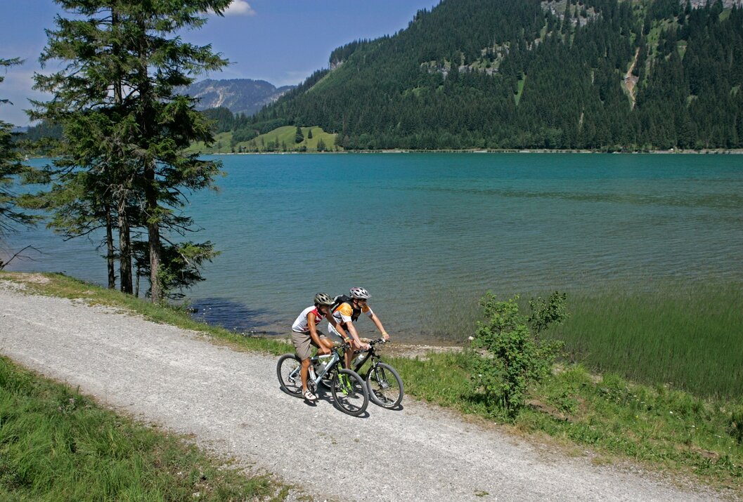 Zwei Radfahrer auf Schotterweg vor blauem See und bewaldeten Bergen