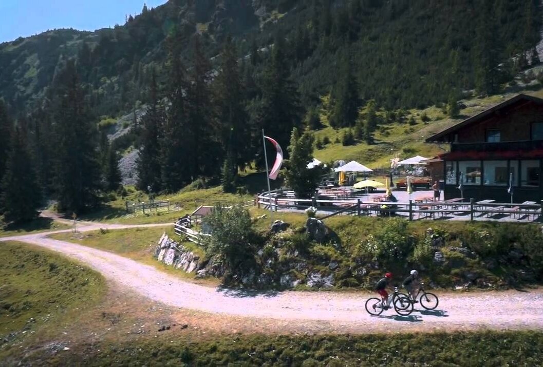 Zwei Mountainbiker fahren auf Schotterweg bergauf, im Hintergrund Hütte mit Terrasse und Berglandschaft