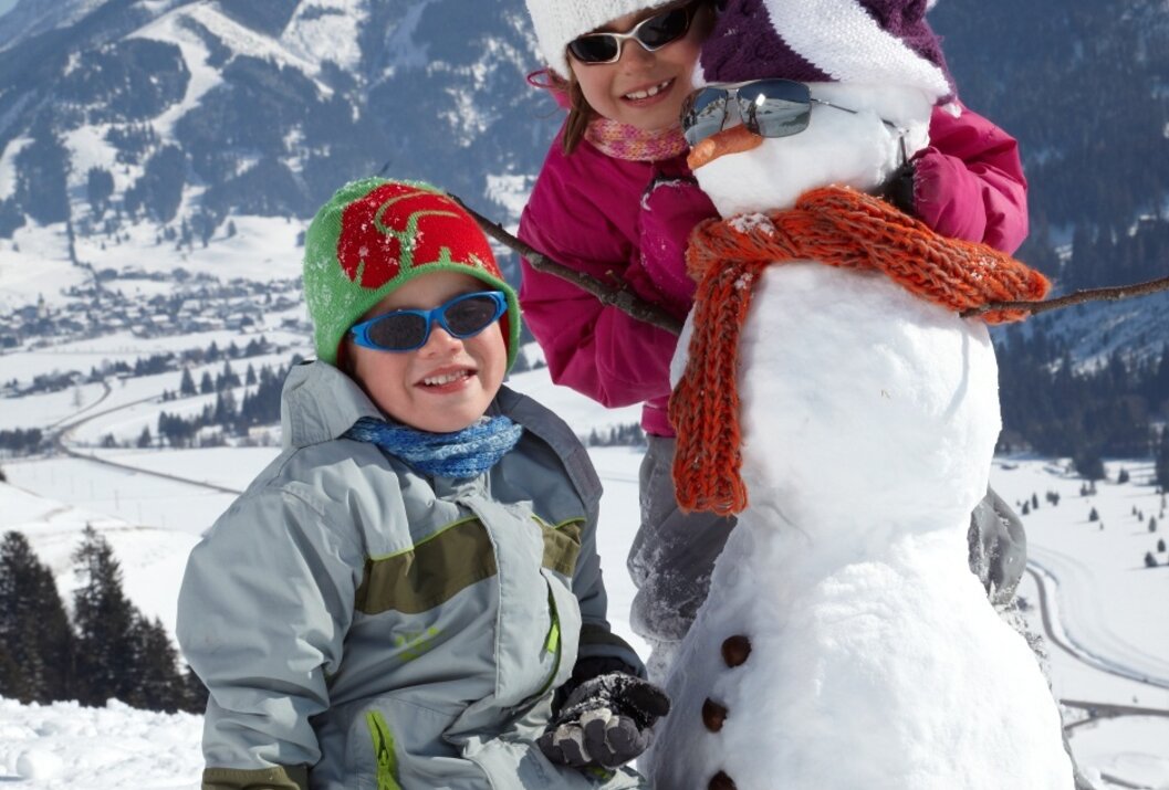Zwei Kinder bauen einen Schneemann vor verschneiter Bergkulisse