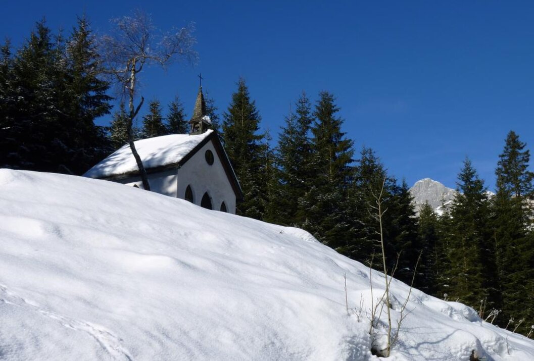 Kleine Kapelle im Schnee vor Tannenwald und blauem Himmel