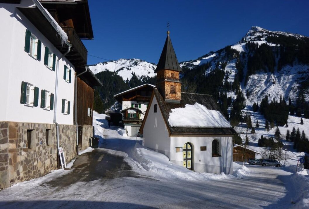 Verschneite Kapelle in Bergdorf vor schneebedeckten Bergen unter blauem Himmel