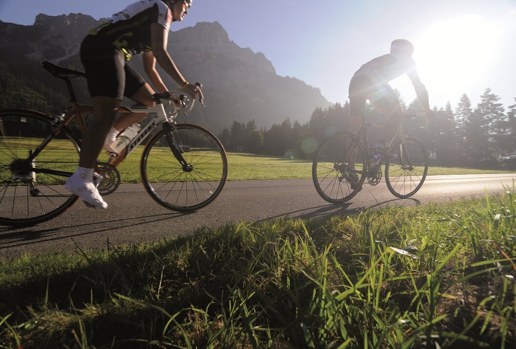 Zwei Radfahrer auf Asphaltstraße vor Bergkulisse und Wiese im Gegenlicht
