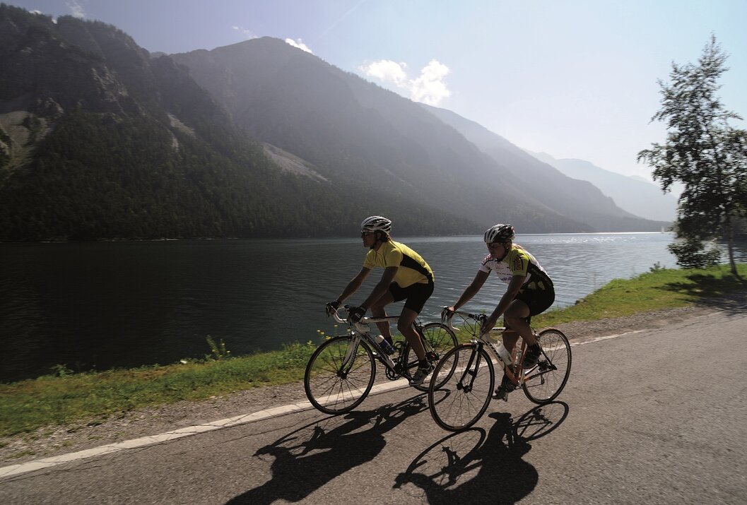 Two cyclists ride on a road in front of a lake and mountains