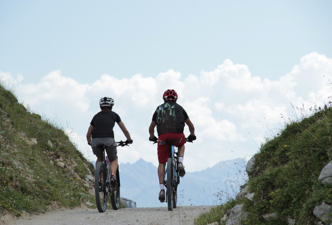 Zwei Mountainbiker fahren bergauf auf einem Schotterweg vor Bergkulisse