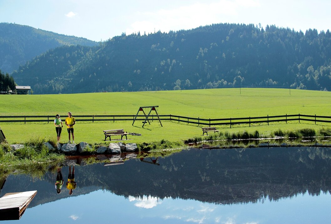 Grüne Wiese mit Spielplatz, umzäunt, spiegelt sich im Wasser, im Hintergrund bewaldete Berge