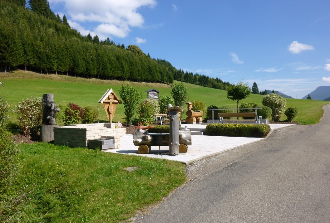 Platz mit Holzkreuz, Bänken und Skulpturen vor grüner Wiese und Wald unter blauem Himmel