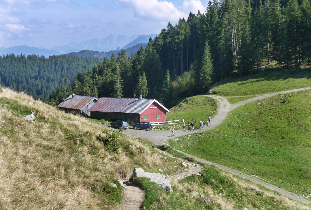 Rote Berghütte auf grüner Wiese, umgeben von Wald und Bergen, Wanderweg führt vorbei