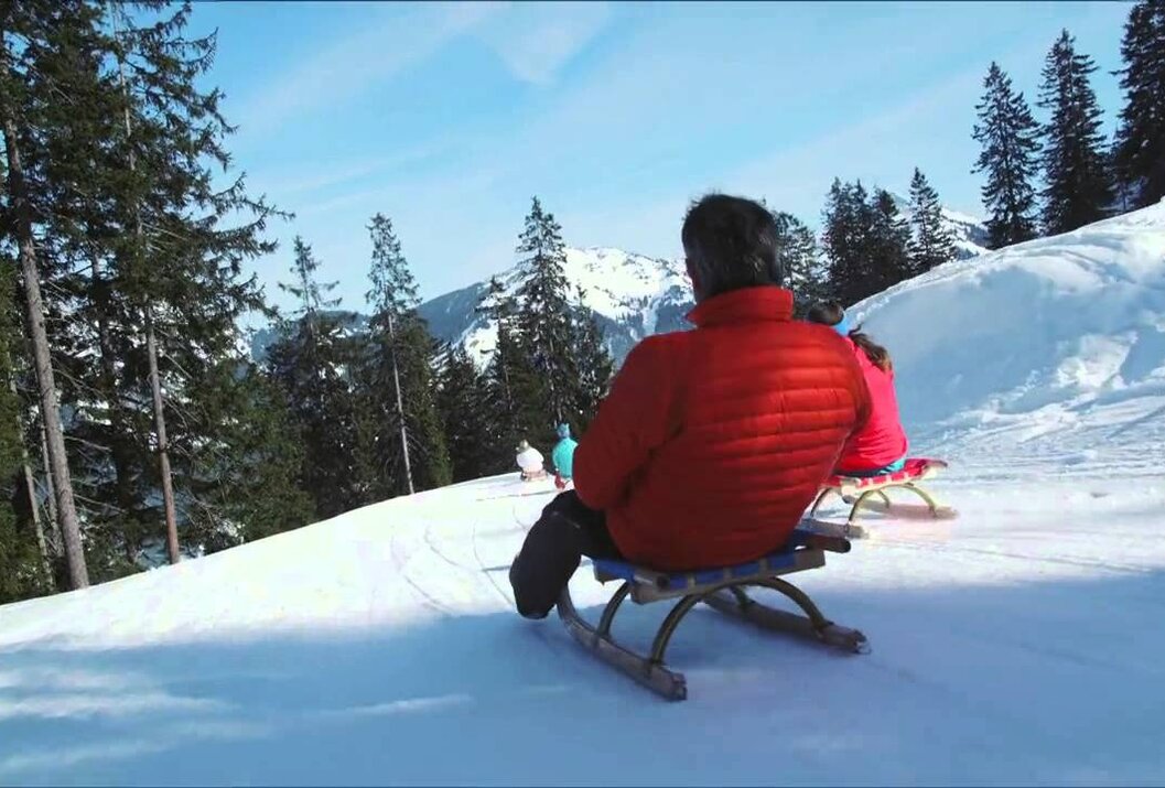 Personen rodeln auf einem schneebedeckten Hügel mit Bäumen und Bergen im Hintergrund