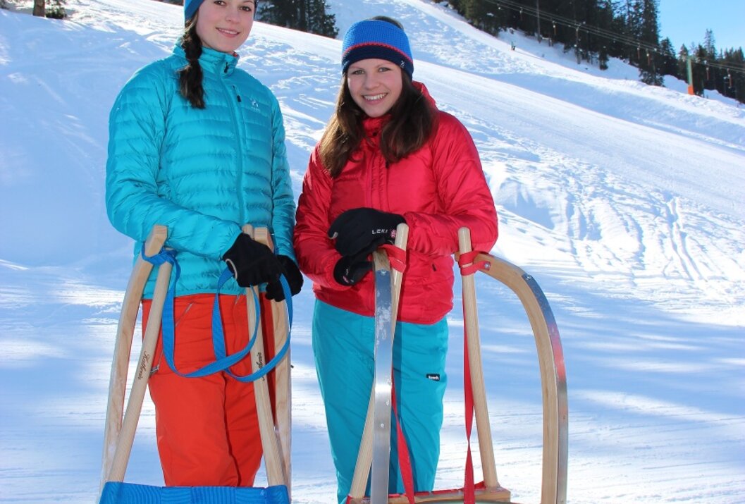 Zwei junge Frauen mit Schlitten im Schnee vor bewaldetem Hang