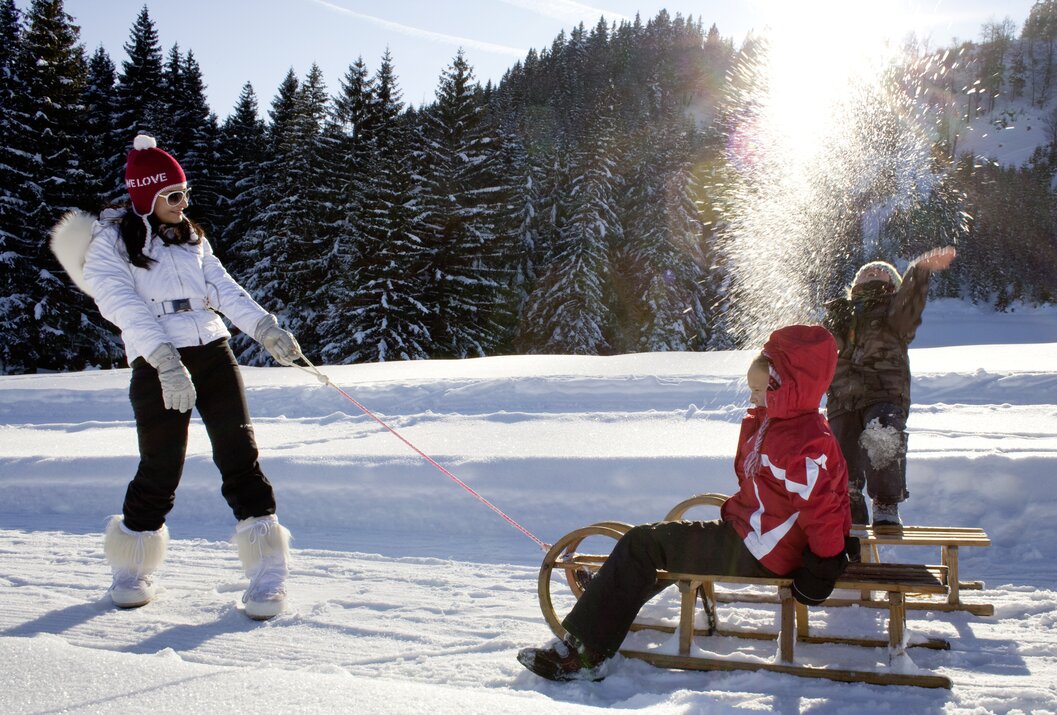 Kind sitzt auf Schlitten, wird von Frau im Schnee gezogen, Wald im Hintergrund