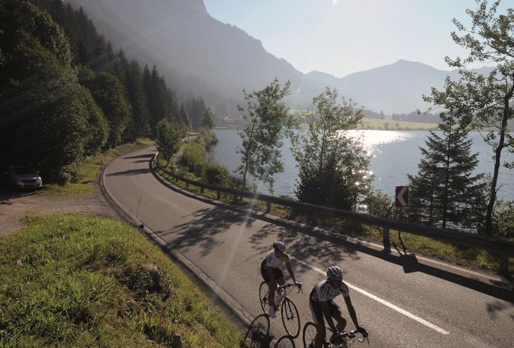 Zwei Radfahrer auf kurviger Straße am See, bewaldete Berge im Hintergrund
