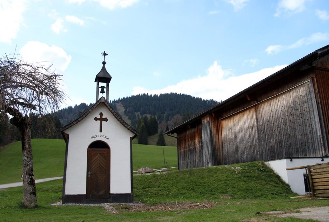 Kleine weiße Kapelle neben einem Holzgebäude vor grüner Wiese und bewaldetem Hügel unter blauem Himmel