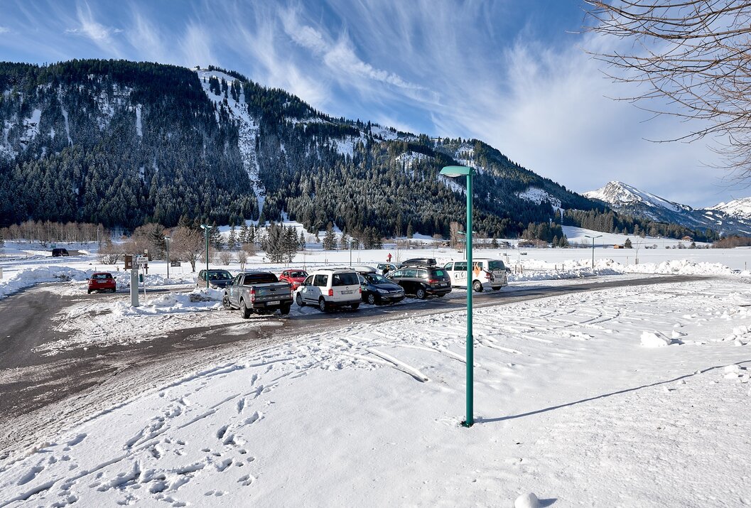 Verschneiter Parkplatz vor bewaldetem Berg unter blauem Himmel