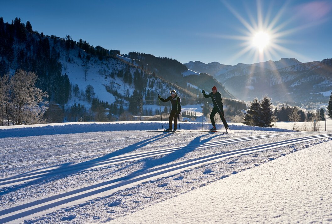 Zwei Langläufer auf Loipe vor Bergpanorama unter strahlender Sonne