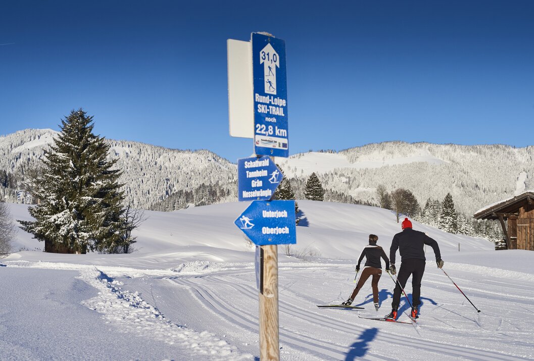 Wegweiser für Skilanglauf vor verschneiter Winterlandschaft mit zwei Langläufern