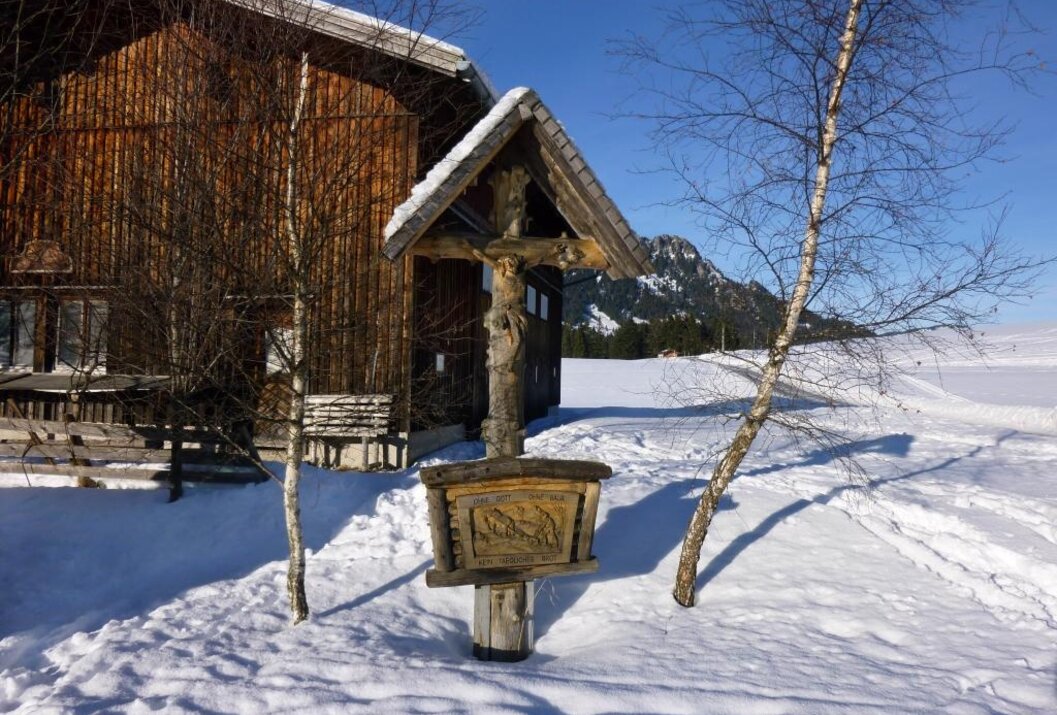 Holzkreuz im Schnee vor Holzhütte und Bergkulisse