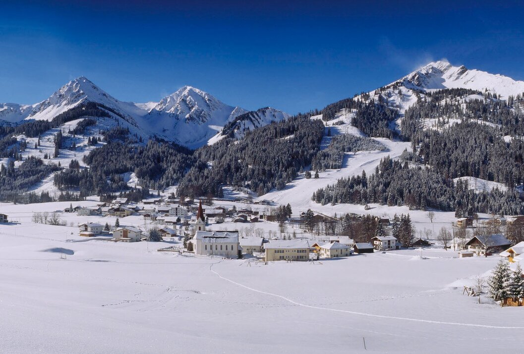 Verschneite Ortschaft mit Kirche vor Bergpanorama unter blauem Himmel