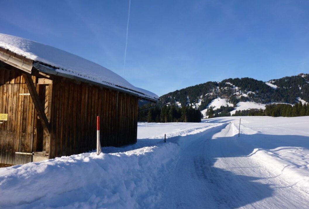 Schneebedeckte Landschaft mit Hütte und Weg vor Bergkulisse unter blauem Himmel