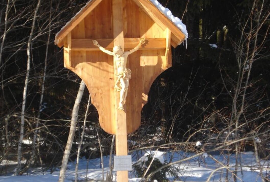 Hölzernes Wegkreuz mit Jesusfigur im Schnee vor dunklem Wald