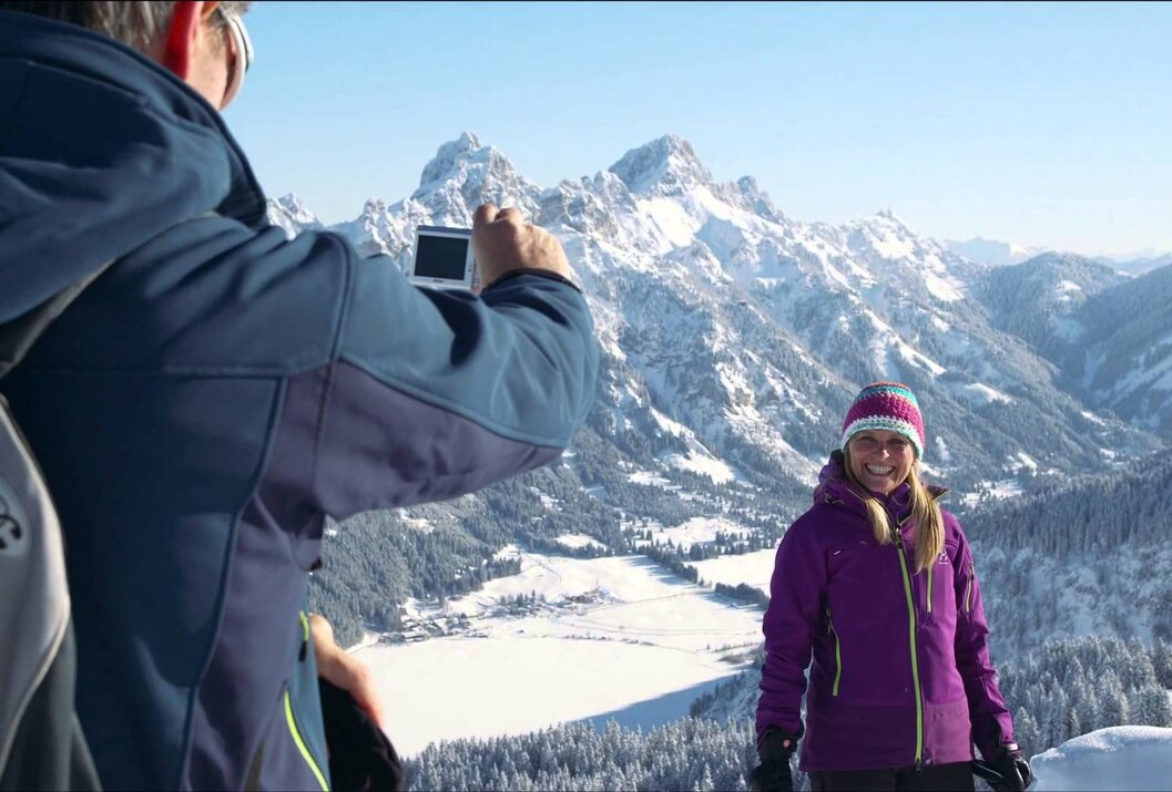 Frau in Winterkleidung vor Bergkulisse, Mann fotografiert sie