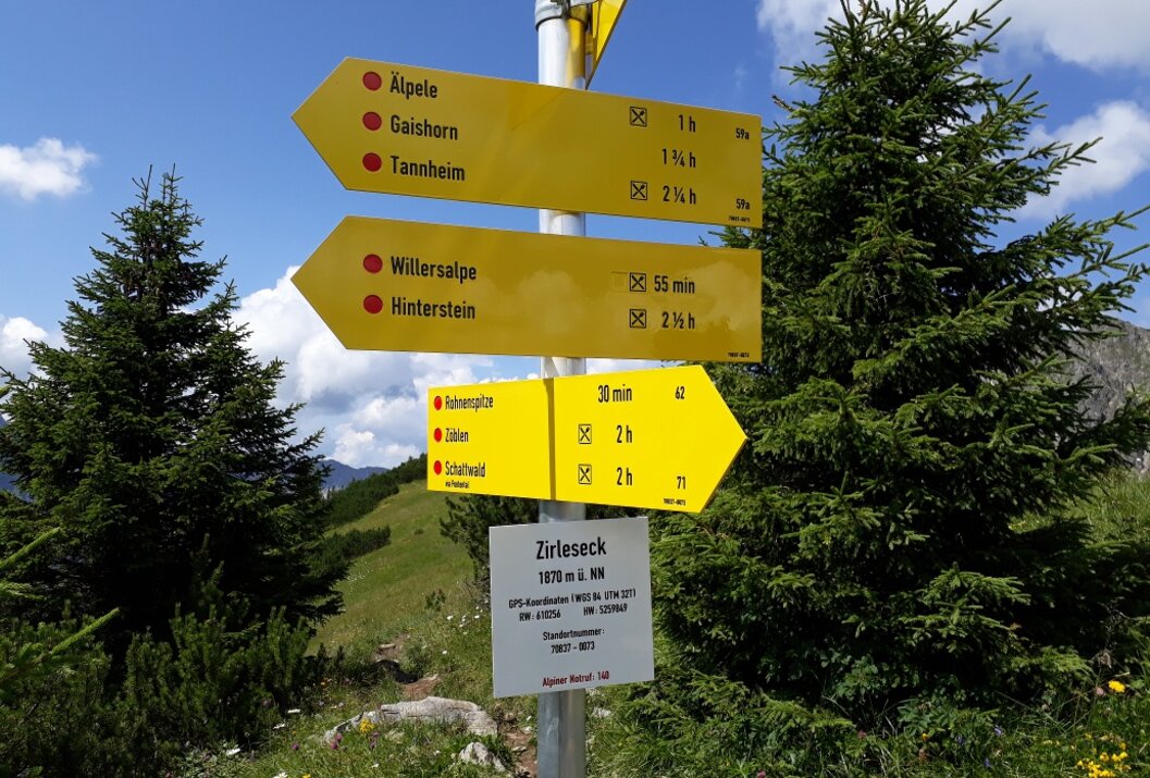 Gelbe Wanderwegweiser mit Ortsnamen und Zeitangaben vor grünen Bäumen