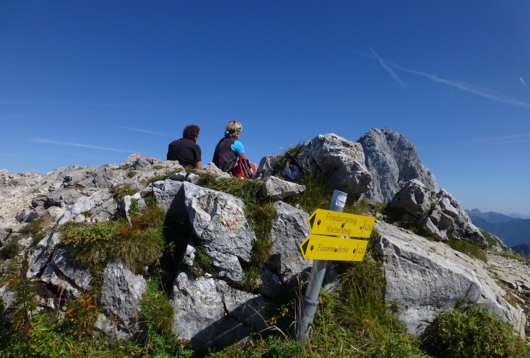 Zwei Personen sitzen auf Felsen mit gelbem Wegweiser vor blauem Himmel