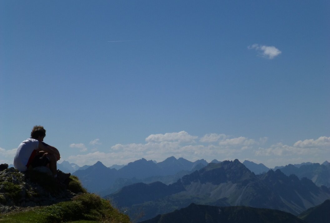Person sitzt auf Bergspitze, Blick auf Bergkette unter blauem Himmel