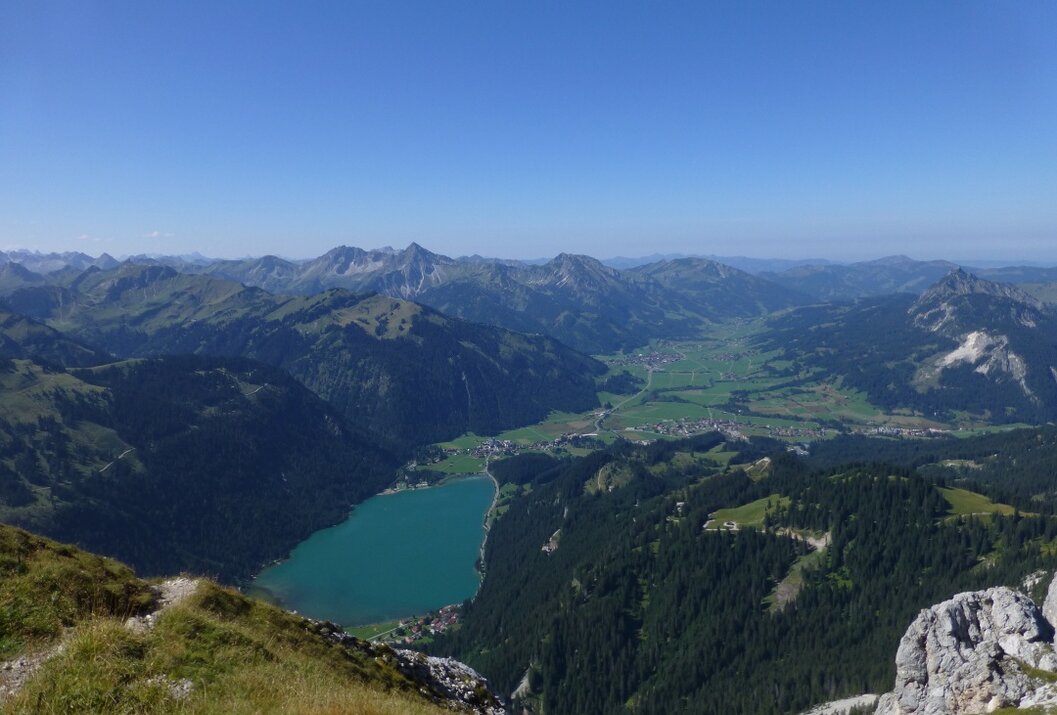 Berglandschaft mit See, grünen Wiesen und bewaldeten Hängen unter blauem Himmel