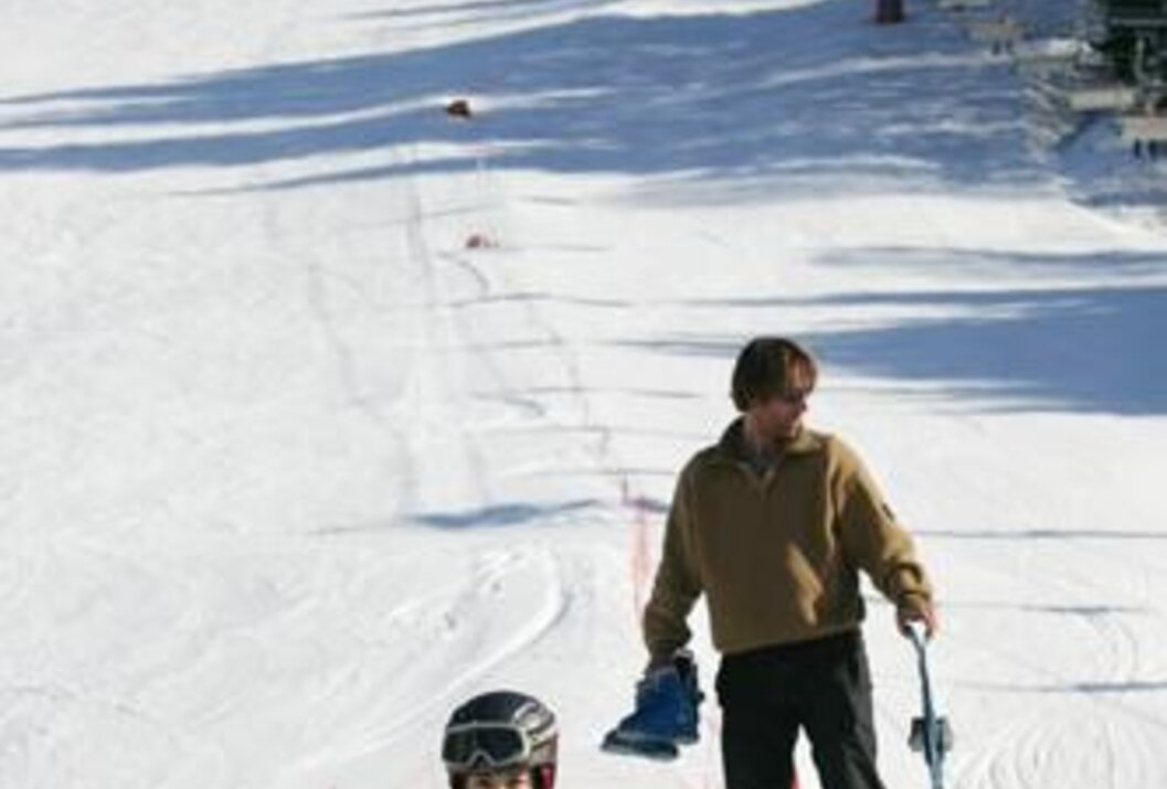 Mann und Kind auf Skipiste, Kind mit Helm, Mann mit Skiern und Skischuhen in der Hand