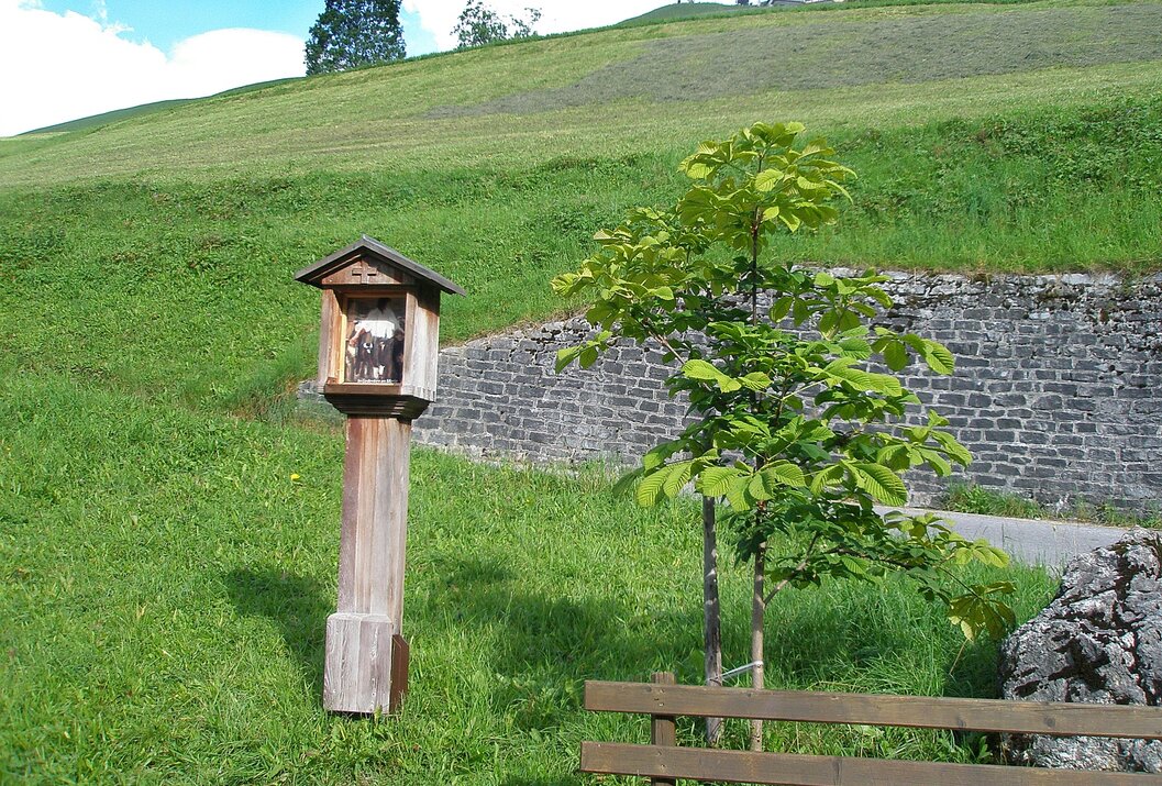 Kleiner Bildstock neben jungem Baum und Bank vor grüner Wiese mit Mauer und Häusern im Hintergrund