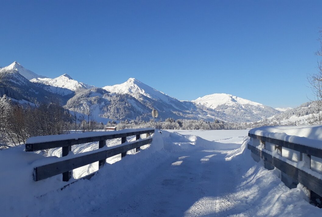 Verschneite Straße mit Geländer vor Bergpanorama unter blauem Himmel