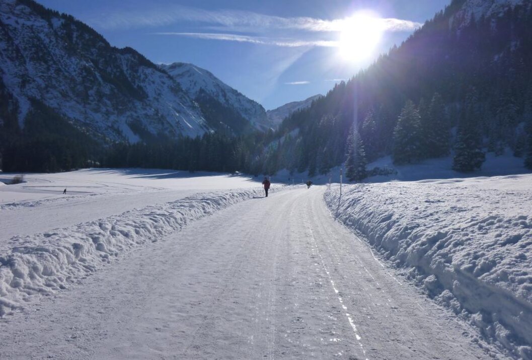 Verschneiter Weg führt durch ein Tal mit Bergen im Hintergrund, die Sonne scheint