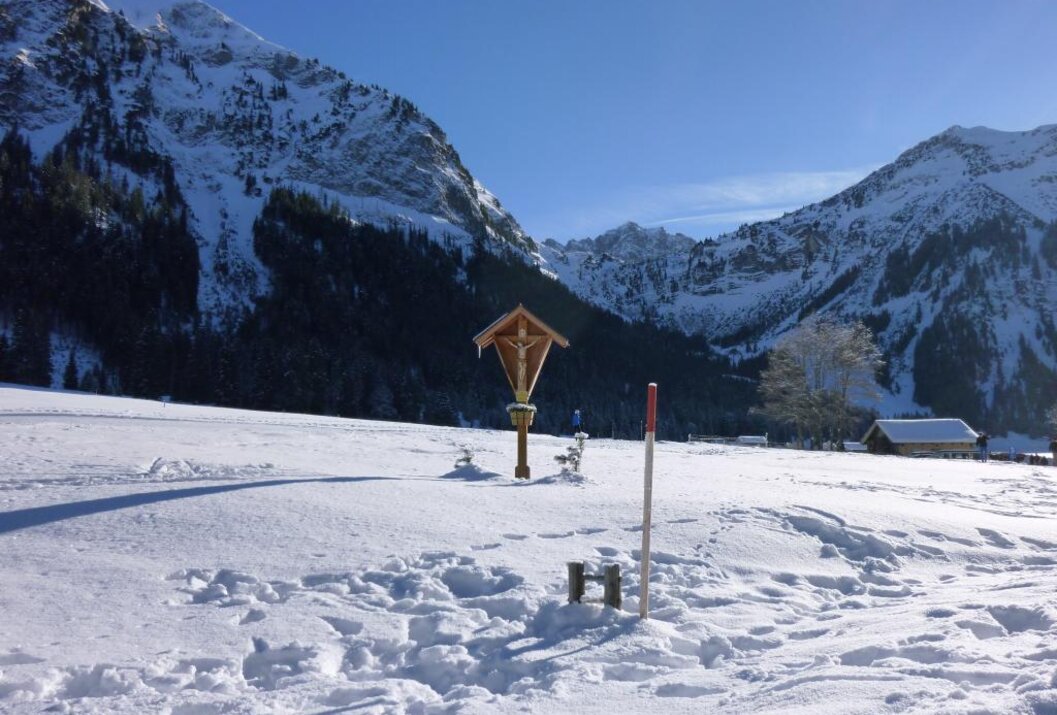 Verschneite Landschaft mit Holzkreuz vor Bergpanorama unter blauem Himmel